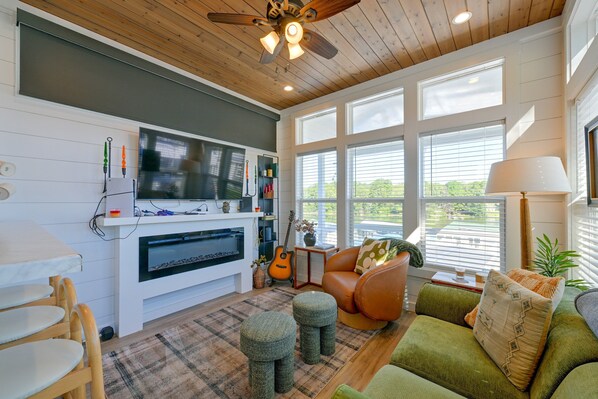 A living room with a fireplace, a television, a guitar, and a large window with blinds. - Lake Norman Views! Tiny Home w/ Deck (Sherrills Ford)