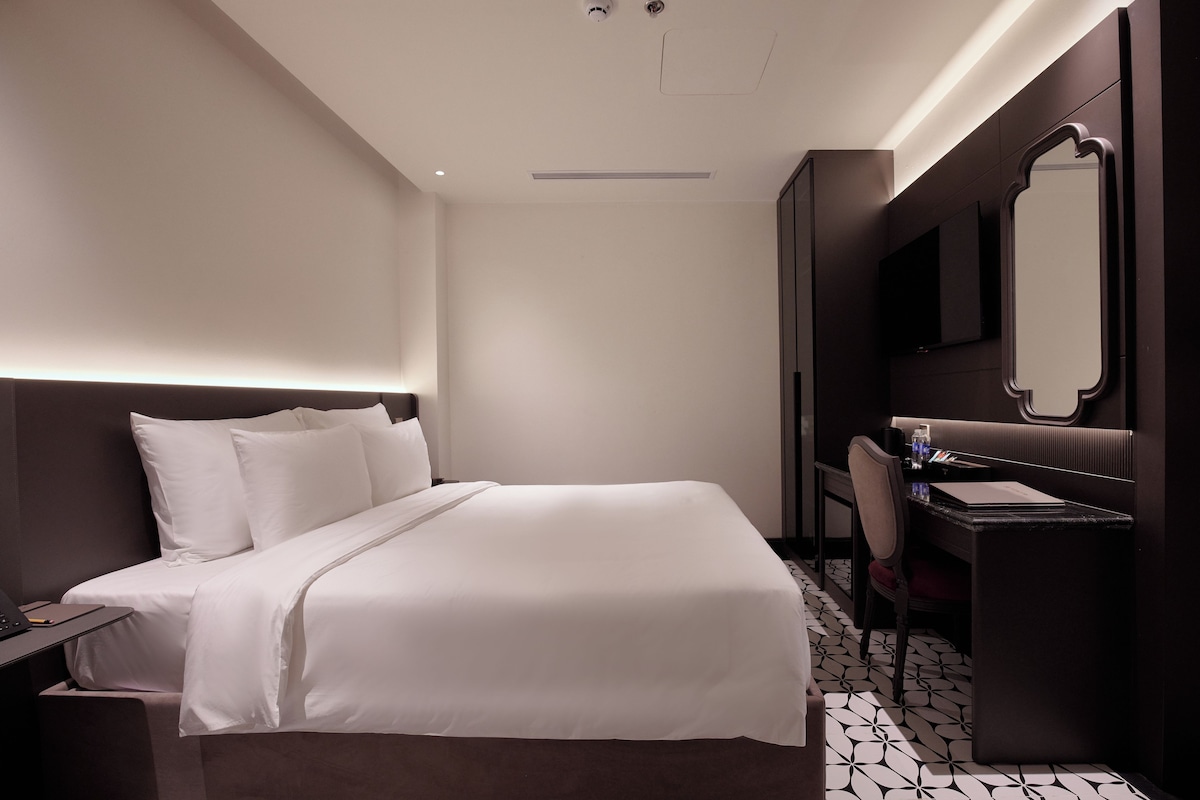 Habitació d'hotel amb un llit gran, una taula de nit, un escriptori amb cadira, un mirall i un paviment amb dibuixos.
