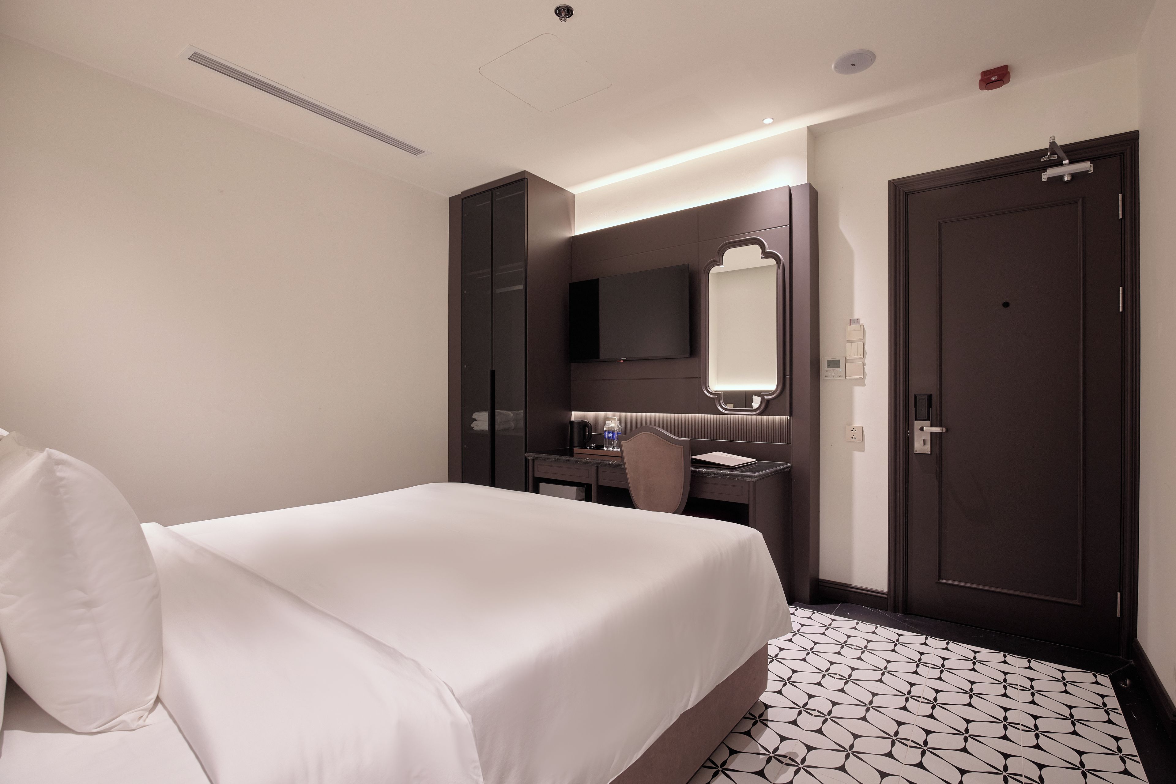 A hotel room with a bed, a desk, a chair, a mirror, and a door.