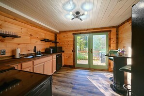 A wooden cabin interior with a kitchen area, a dining table, and a sliding glass door leading to an outdoor deck. - Hocking Hills Area Retreat: Relax & Explore! (Rockbridge)