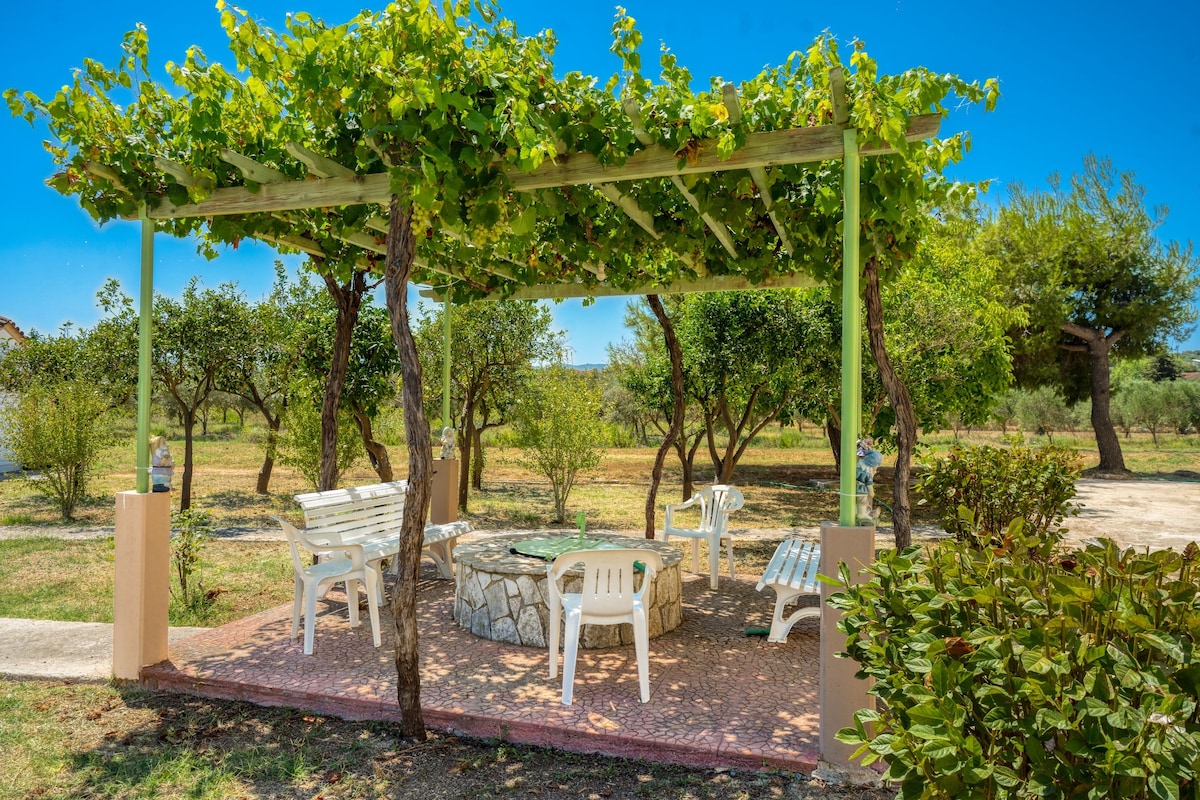 A pergola with a stone table and chairs, surrounded by greenery and trees.