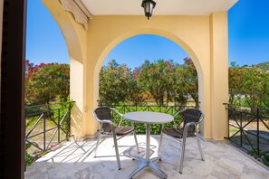 A covered patio area with a table and two chairs, surrounded by a metal railing, overlooking a garden with flowering plants. - Archontiko Studios (Zakynthos)