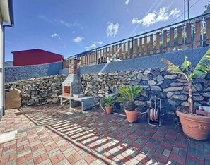 Eine Terrasse mit Steinmauer, Blumentöpfen, Grill und rotem Sonnensegel.