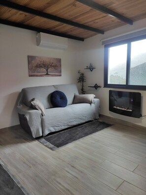 Ein Wohnzimmer mit grauem Sofa, einem Baumgemälde, einem an der Wand befestigten Fernseher und einem Fenster mit Blick.