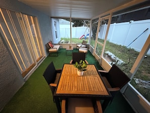 Ein überdachter Patio-Bereich mit Holztisch und Stühlen, einem Blumentopf und Blick auf den Garten.