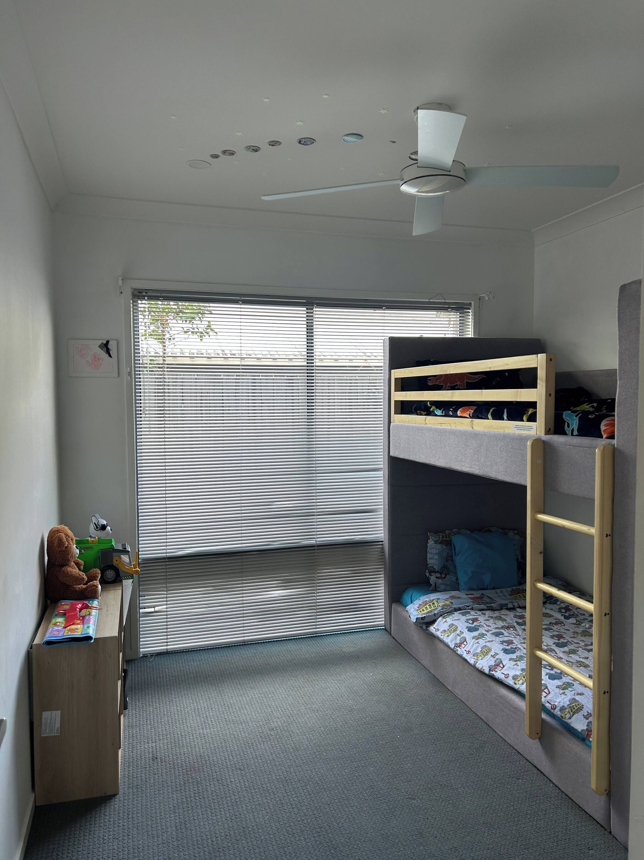 Een kinderkamer met stapelbedden, een bureau, een stoel en een raam met jaloezieën.