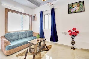 Ein Wohnzimmer mit einer Couch, einem kleinen Tisch, einer Vase mit Blumen, einem Fenster mit Vorhängen und einem an der Wand hängenden Bild.