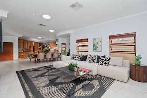 Ein modernes Wohnzimmer mit einem Ecksofa, einem Couchtisch und einem Essbereich, im Hintergrund ist eine Küche zu sehen.