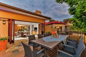 An outdoor dining area with a table and chairs, a view of the interior, and a red-roofed building in the background. - Luxurious 4-Bedroom Villa with Private Pool in Hartbeespoort (Hartbeespoort)