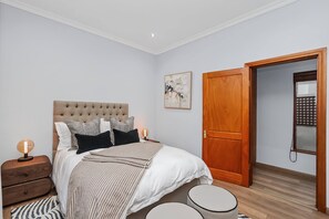 Ein Schlafzimmer mit einem gesteppten Kopfteil, einem Bett mit gemusterten Betttextilien, einem Nachttisch mit Lampe, einer Holztür und einem an der Wand befestigten Kunstwerk.