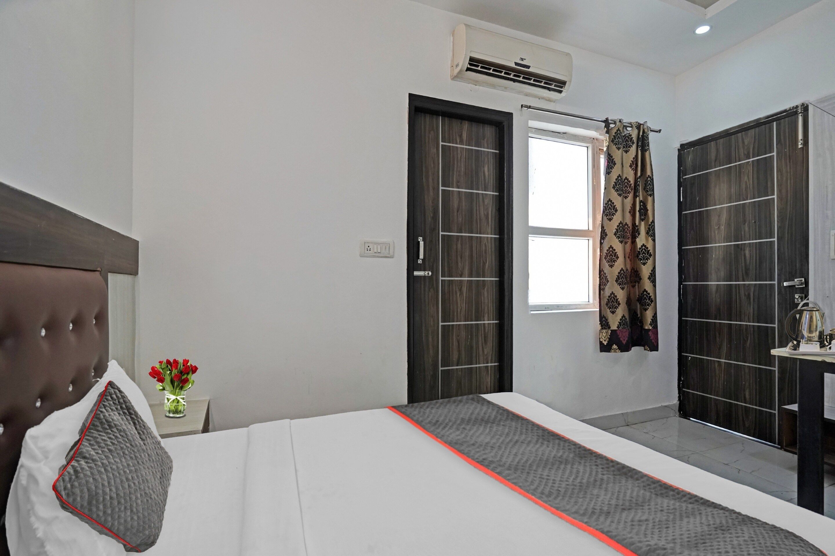 Une chambre d’hôtel comprenant un lit, une fenêtre avec des rideaux, un appareil de climatisation, une table de chevet avec une bouilloire et une armoire fixée au mur.
