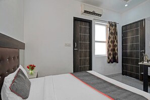 A hotel room with a bed, a window with curtains, an air conditioning unit, a bedside table with a kettle, and a wall-mounted wardrobe. - Signature Hotel Noida-62 (Noida)