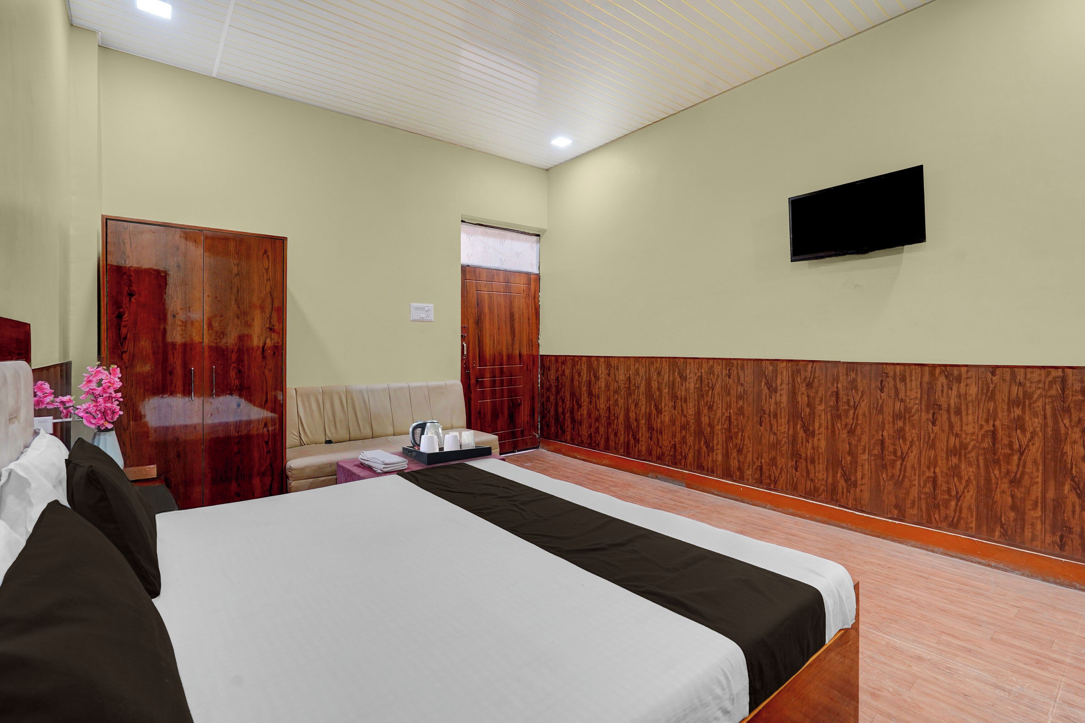 A hotel room with a bed, a sofa, a wooden wardrobe, a television mounted on the wall, and a window with curtains.