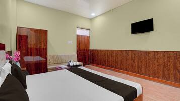 A hotel room with a bed, a sofa, a wooden wardrobe, a television mounted on the wall, and a window with curtains.