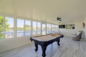A pool table in a room with large windows overlooking a body of water, a ceiling fan, and a wicker chair. - Right on Canal! Sleek Home Near Gulf Coast Fun (Hudson)