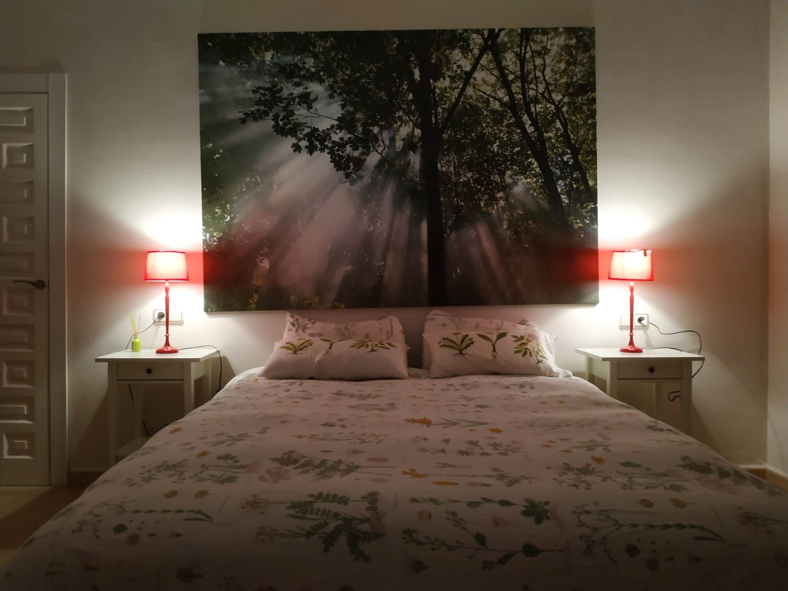 Un dormitorio con una cama, dos mesitas de noche, dos lámparas y un cuadro grande en la pared de un árbol con luz solar.