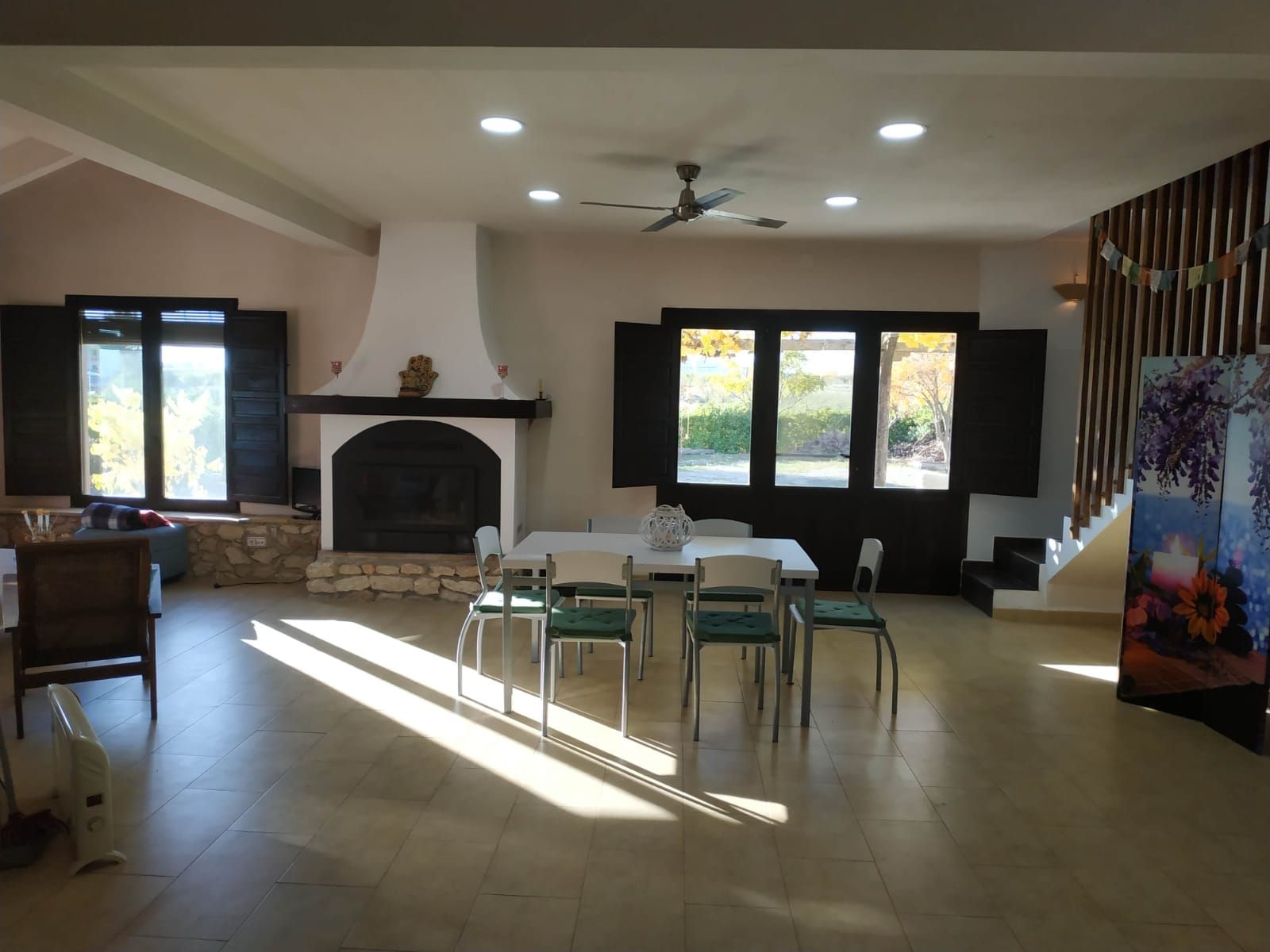 A spacious living room with a fireplace, a dining area with chairs, and large windows with shutters.