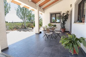 Un patio techado con mesa y sillas, rodeado de plantas en macetas y vistas al exterior.