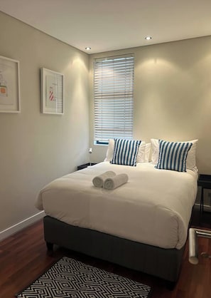 A neatly made bed with pillows, a bedside table, and a window with blinds. - Manhattan Feeling! 2BR, Huge Terrace & Epic Views, 2min walk Bree Street (Cape Town)