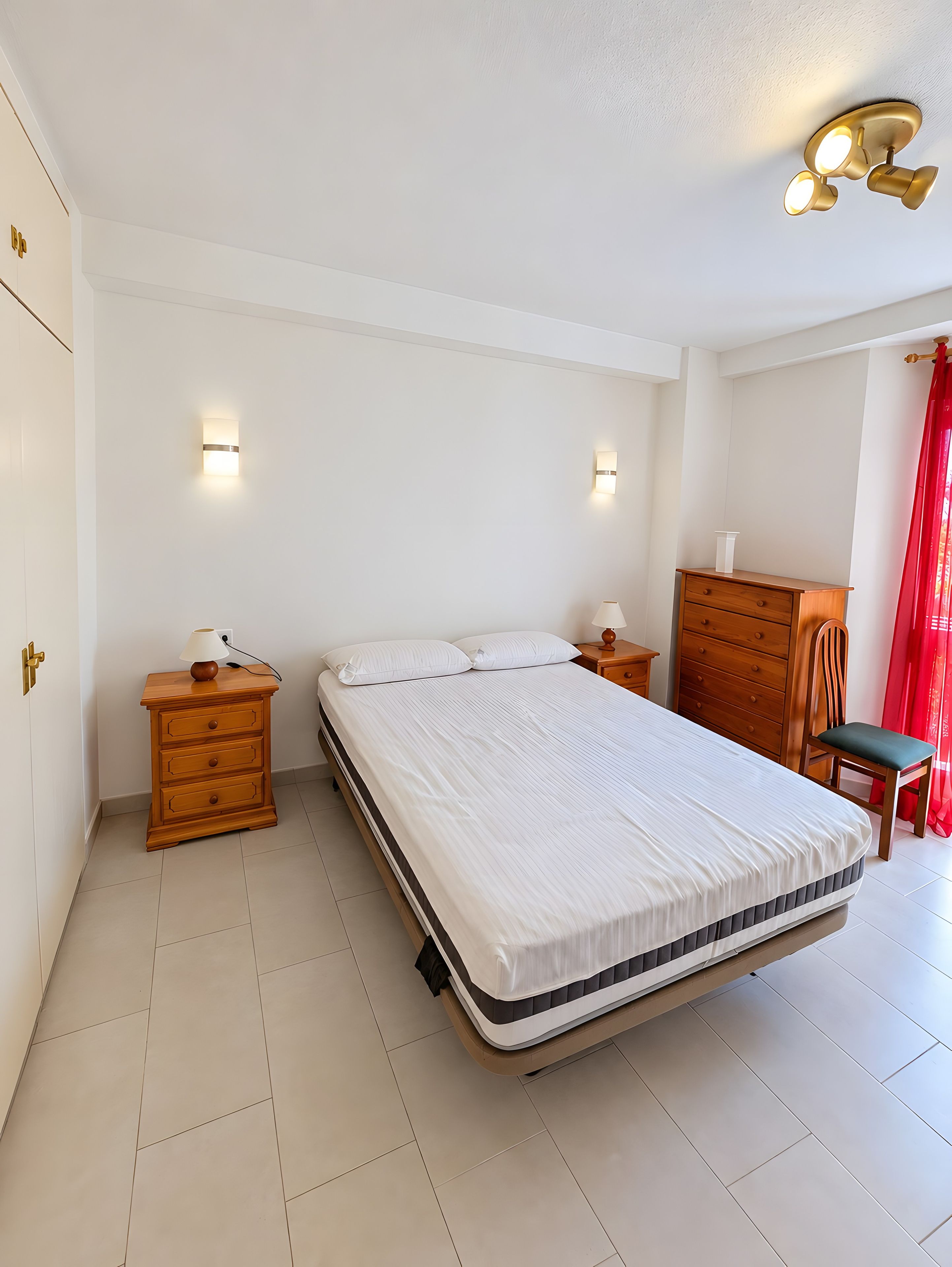 A bedroom with a bed, wooden nightstands, a dresser, and wall sconces.