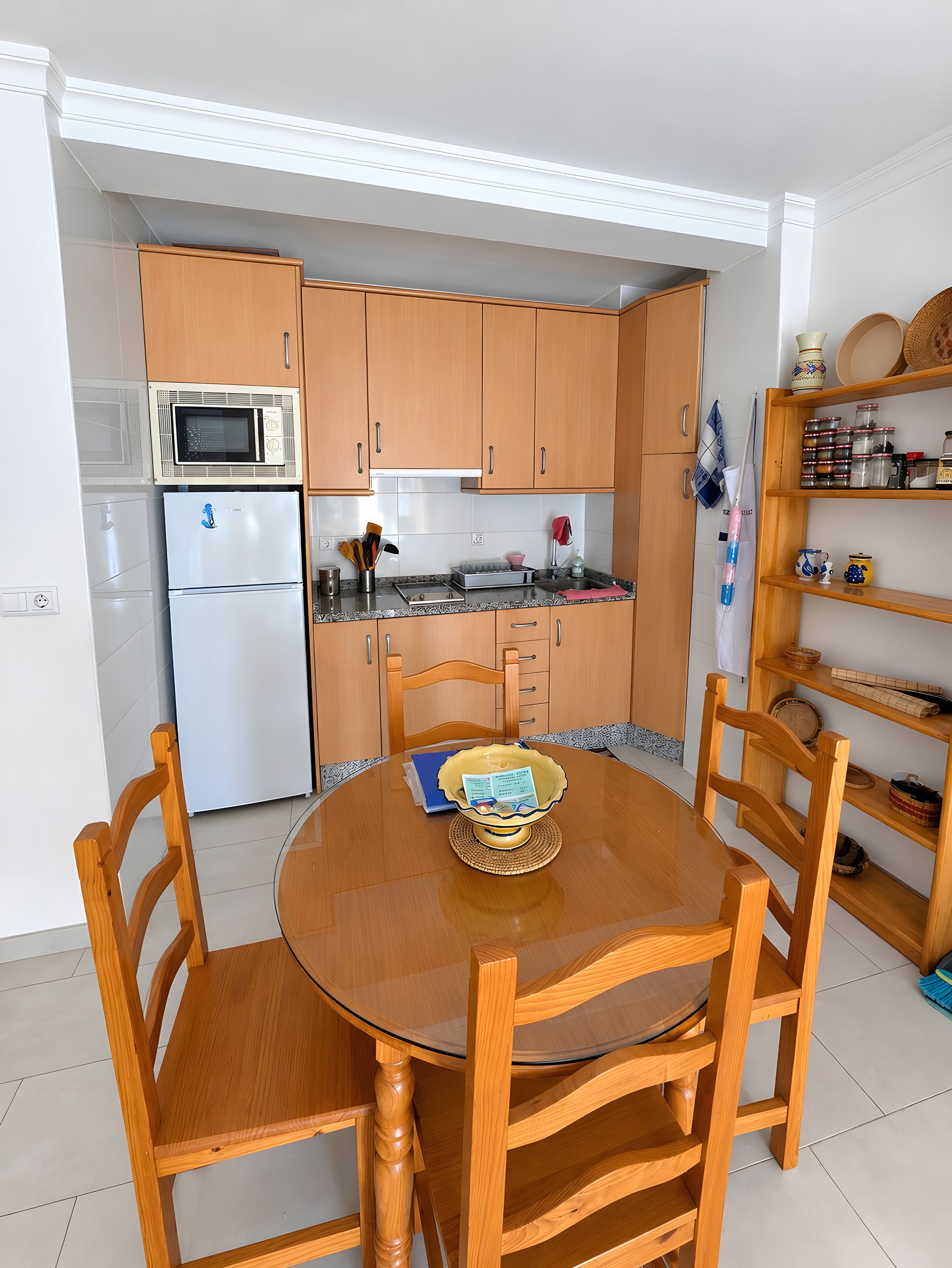 Een compacte keuken met houten kastjes, een witte koelkast, een magnetron en een eettafel met stoelen.