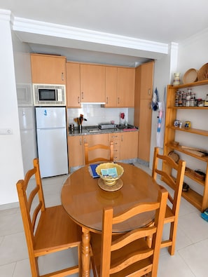 Een compacte keuken met houten kastjes, een witte koelkast, een magnetron en een eettafel met stoelen.