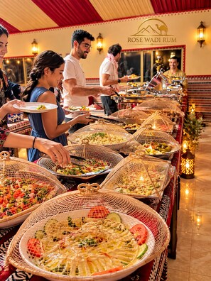 A buffet setup with various dishes in woven baskets, a sign reading 'ROSE WADI RUM', and a warm interior ambiance. - Rose Wadi Rum Luxury Bubble Camp (Wadi Rum Village)