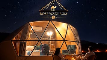 Une structure en forme de dôme géodésique offrant une vue sur le ciel nocturne, un logo représentant un chameau et l’inscription « ROSE WADI RUM », et deux personnes assises à l’intérieur.
