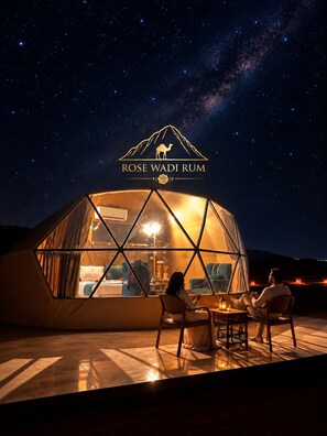 A geodesic dome structure with a view of the night sky, a logo with a camel and the text 'ROSE WADI RUM', and two people seated inside. - Rose Wadi Rum Luxury Bubble Camp (Wadi Rum Village)