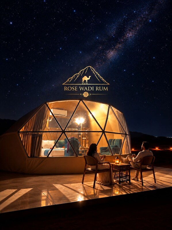 A geodesic dome structure with a view of the night sky, a logo with a camel and the text 'ROSE WADI RUM', and two people seated inside.