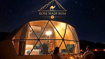 En geodisk kupolkonstruktion med utsikt över natthimlen, en logotyp med en kamel och texten ”ROSE WADI RUM”, och två personer som sitter inuti.