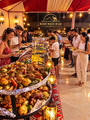 A buffet with a variety of dishes, a sign reading 'ROSE WADI RUM', and people serving themselves.