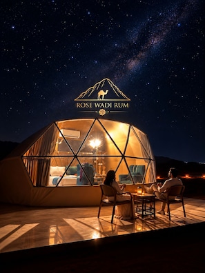 A geodesic dome structure with a view of the night sky, a logo with a camel and the text 'ROSE WADI RUM', and two people seated inside.