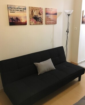 En svart soffa med en enda kudde, tre inramade tavlor på väggen och en golvlampa.