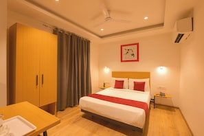 A hotel room with a bed, a wooden wardrobe, a desk with a tray, a ceiling fan, a wall-mounted artwork, and a bedside table with a lamp. - Hotel Capital Leaf (New Delhi)