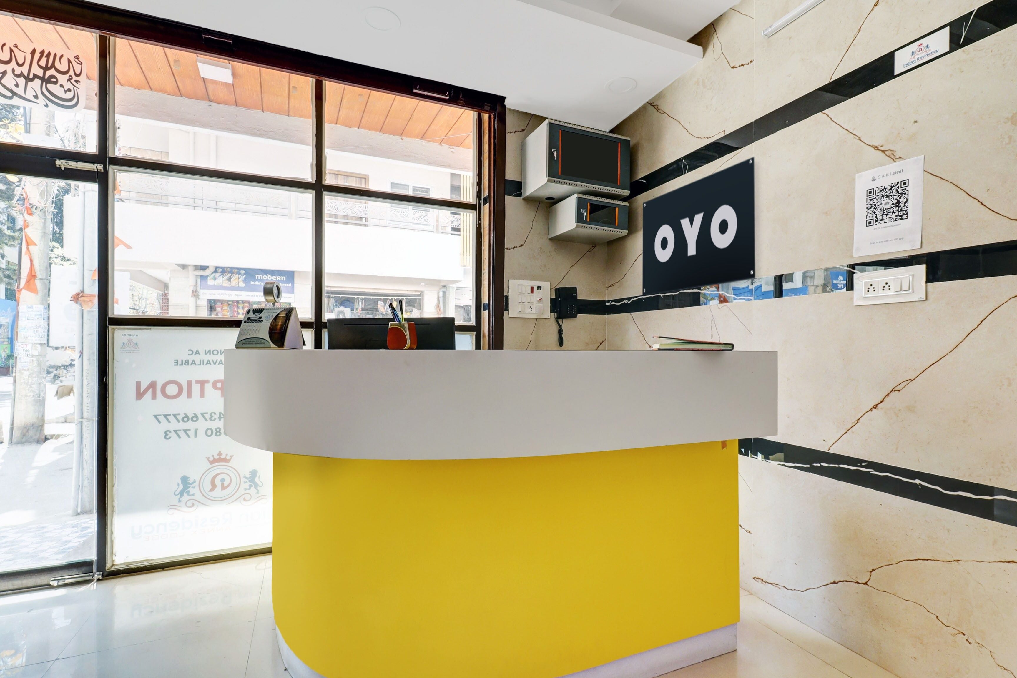 A reception desk with a yellow base and a white top, a wall-mounted air conditioning unit, and a sign with 'OYO' branding.