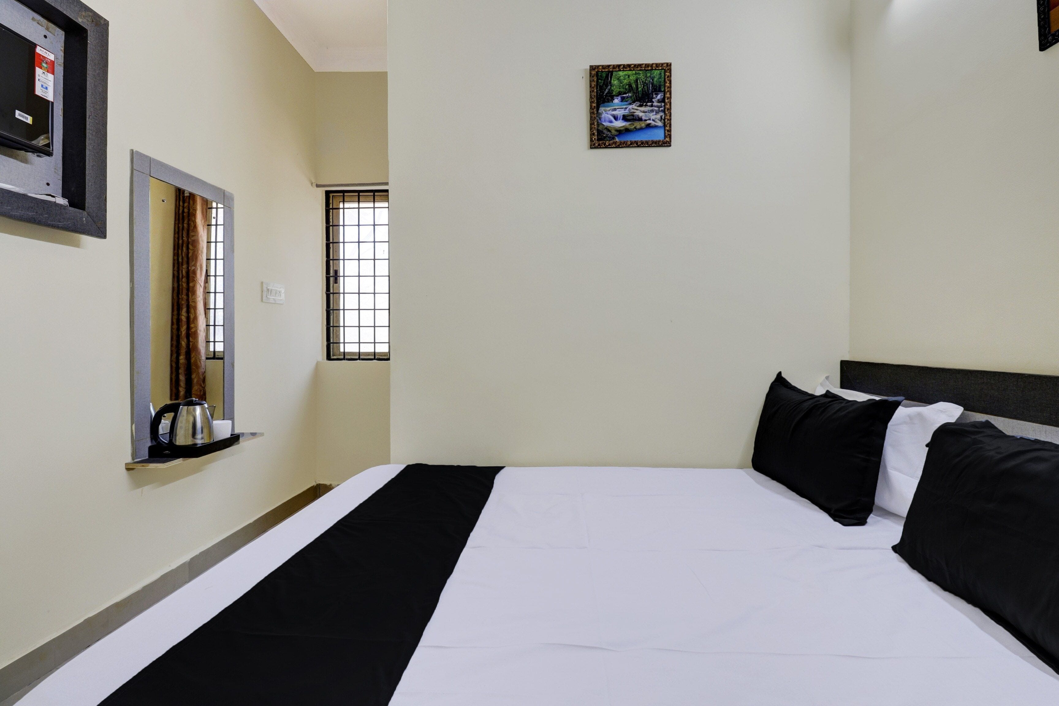 A hotel room with a bed, a window, a wall-mounted picture, and a small shelf with a kettle.