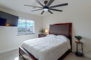Ein Schlafzimmer mit einem Bett, einem Deckenventilator, einem Fenster, einem Nachttisch, einer Lampe und einem Blumentopf.