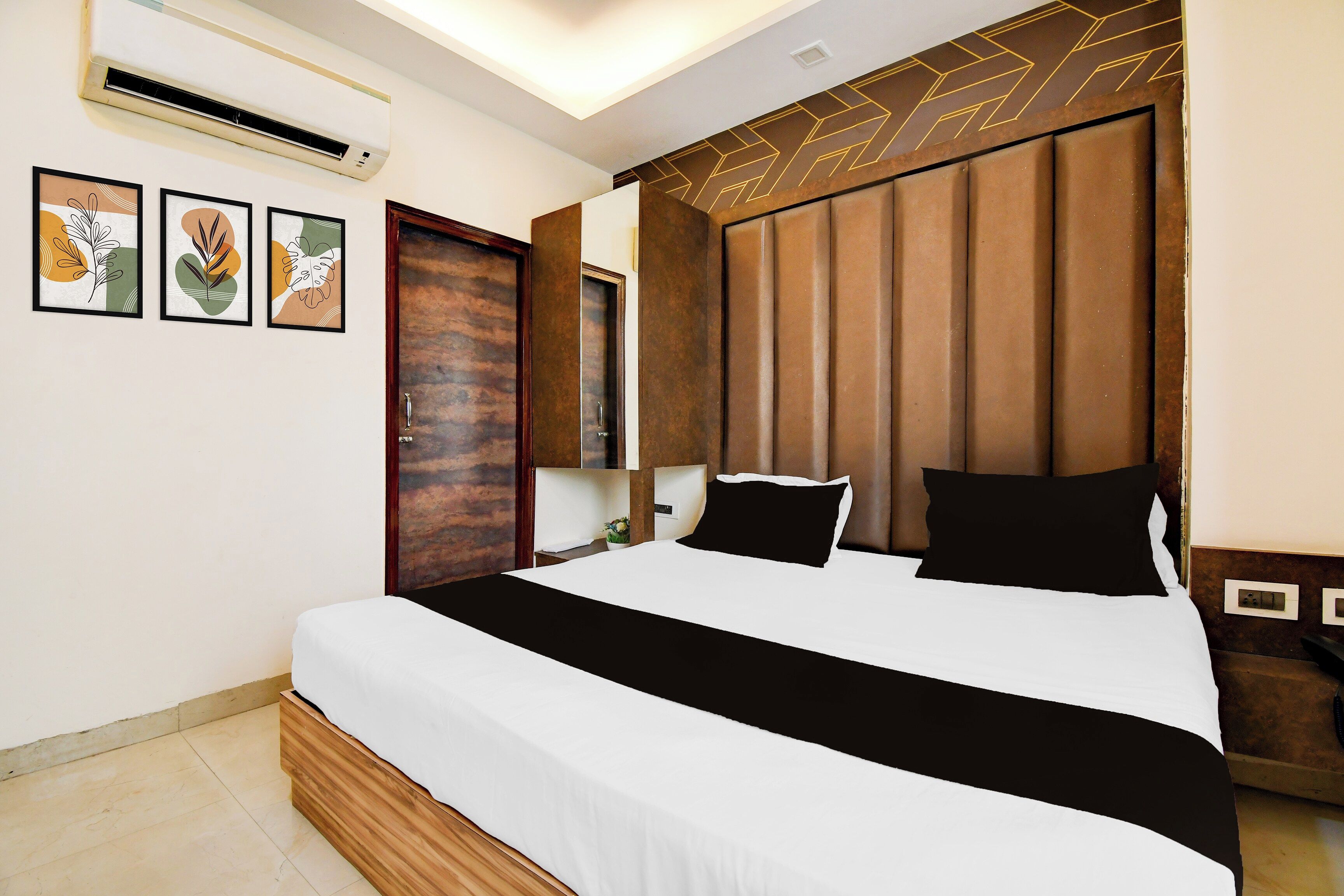 Una habitación de hotel con una cama grande, cabecero de madera, aire acondicionado de pared y tres cuadros enmarcados.