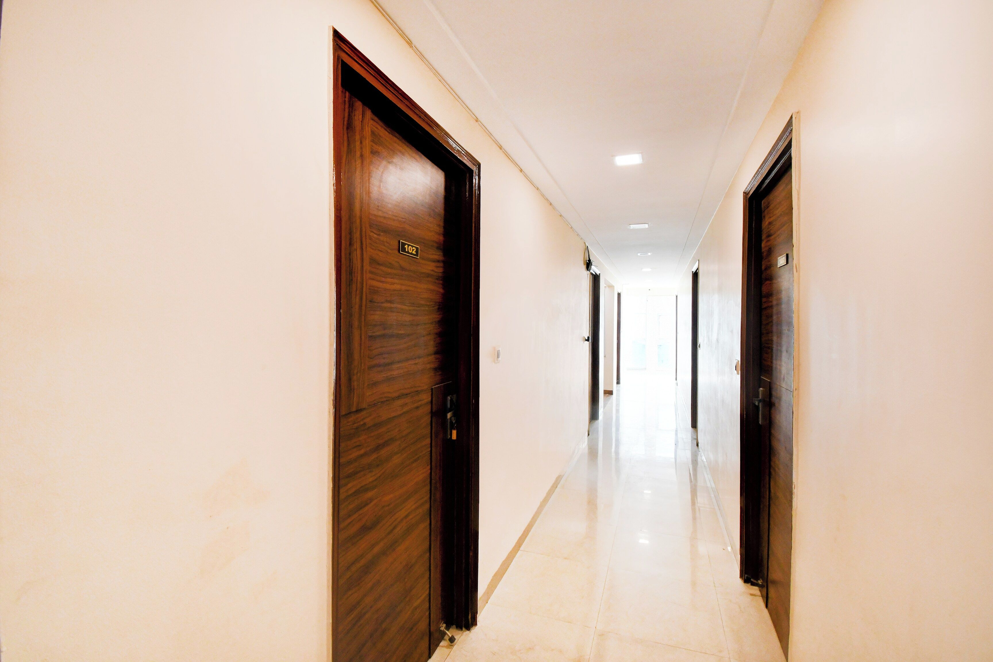A corridor with wooden doors, each marked with a number. The corridor has a tiled floor and is well-lit with ceiling lights.