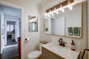 A bathroom with a white toilet, a vanity with a marble countertop, a large mirror with lights, and a view of a hallway. - Pet-Friendly Central Home w/ Fenced Yard! (San Diego)