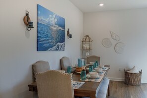 Uma área de refeições com mesa de madeira posta para quatro pessoas, cadeiras, luminária fixada na parede e uma pintura de uma paisagem costeira.