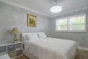 Ein Schlafzimmer mit Bett, Nachttisch, Lampe, einem Gemälde und einem Fenster mit Jalousien.