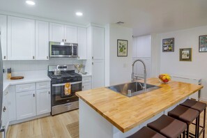 A modern kitchen with white cabinets, stainless steel appliances, a wooden island with bar stools, and a fruit bowl. - North Hollywood Home, Extended Stay (Los Angeles)