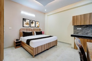 Quarto de hotel com cama, mesa de cabeceira e pequena escrivaninha com cadeira. Quadros emoldurados decoram a parede e um armário de madeira possui bancada de mármore.