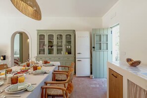 A dining area with a long table set for breakfast, a cabinet with glass doors, a refrigerator, and a window with a view. - Monte do Areal (Santiago do cacem)