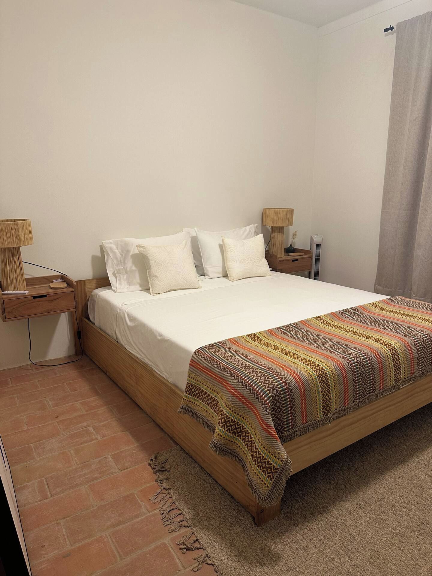 A single-bed room with a wooden headboard, a bedside table with a lamp, a bedside plant, and a patterned throw blanket.