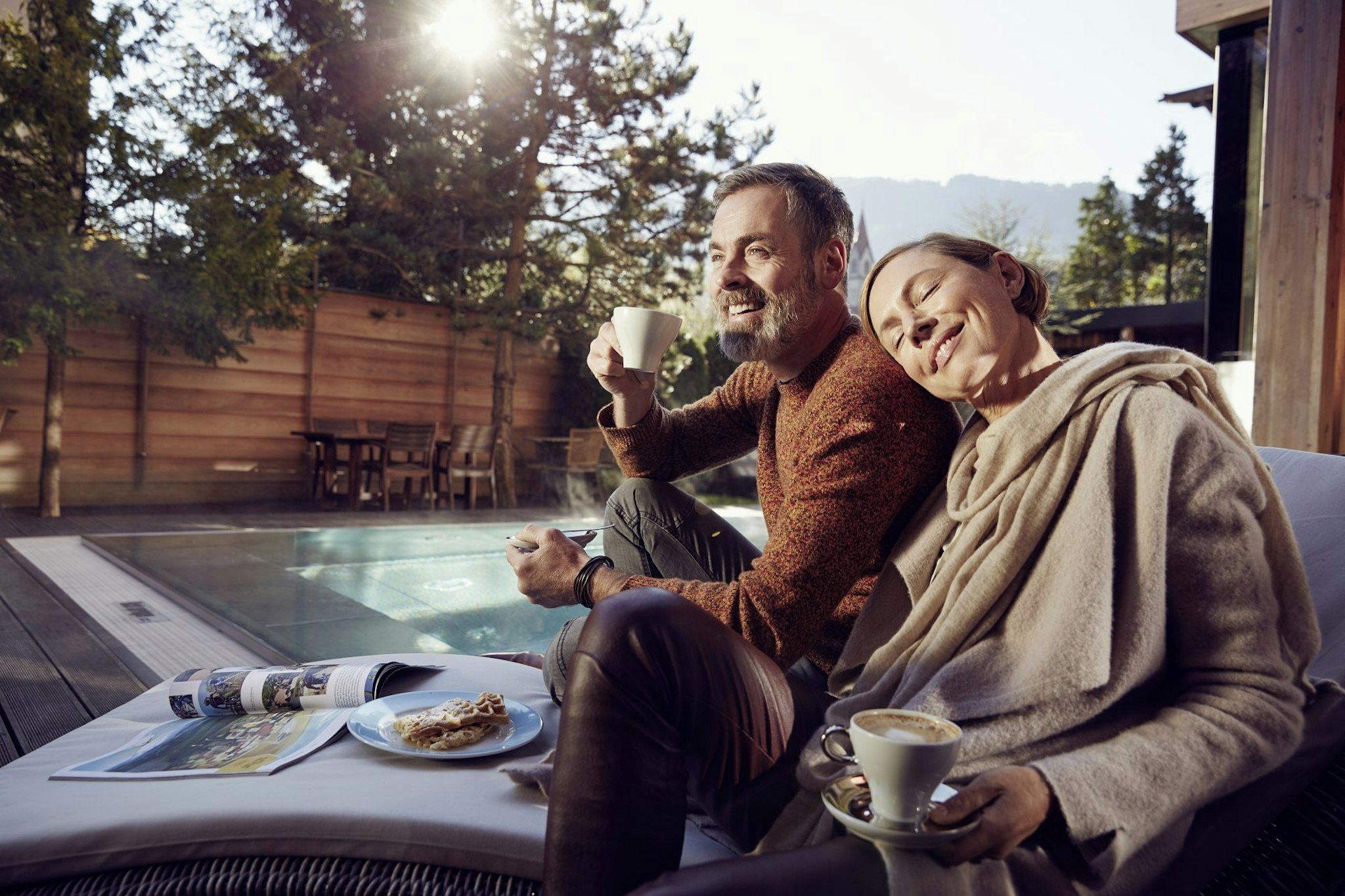 Un couple est assis au bord d’une piscine, une table se trouve devant eux, sur laquelle sont posés un journal et une assiette de nourriture.