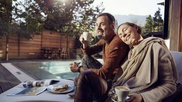Un couple est assis au bord d’une piscine, une table se trouve devant eux, sur laquelle sont posés un journal et une assiette de nourriture.