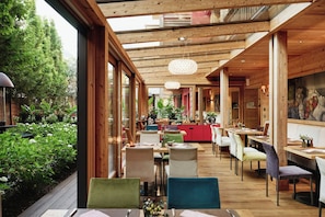 Ein Restaurant mit Holzbalken, großen Fenstern und Blick auf Grünflächen. Tische und Stühle sind zum Essen bereitgestellt.
